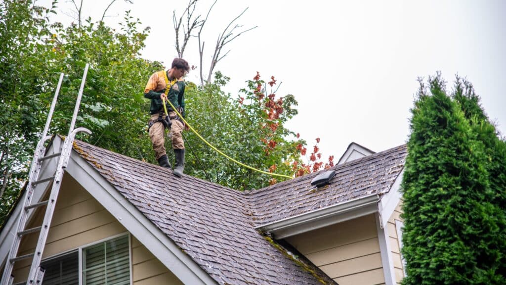 House roof being cleaned by a man using hose and water spray.