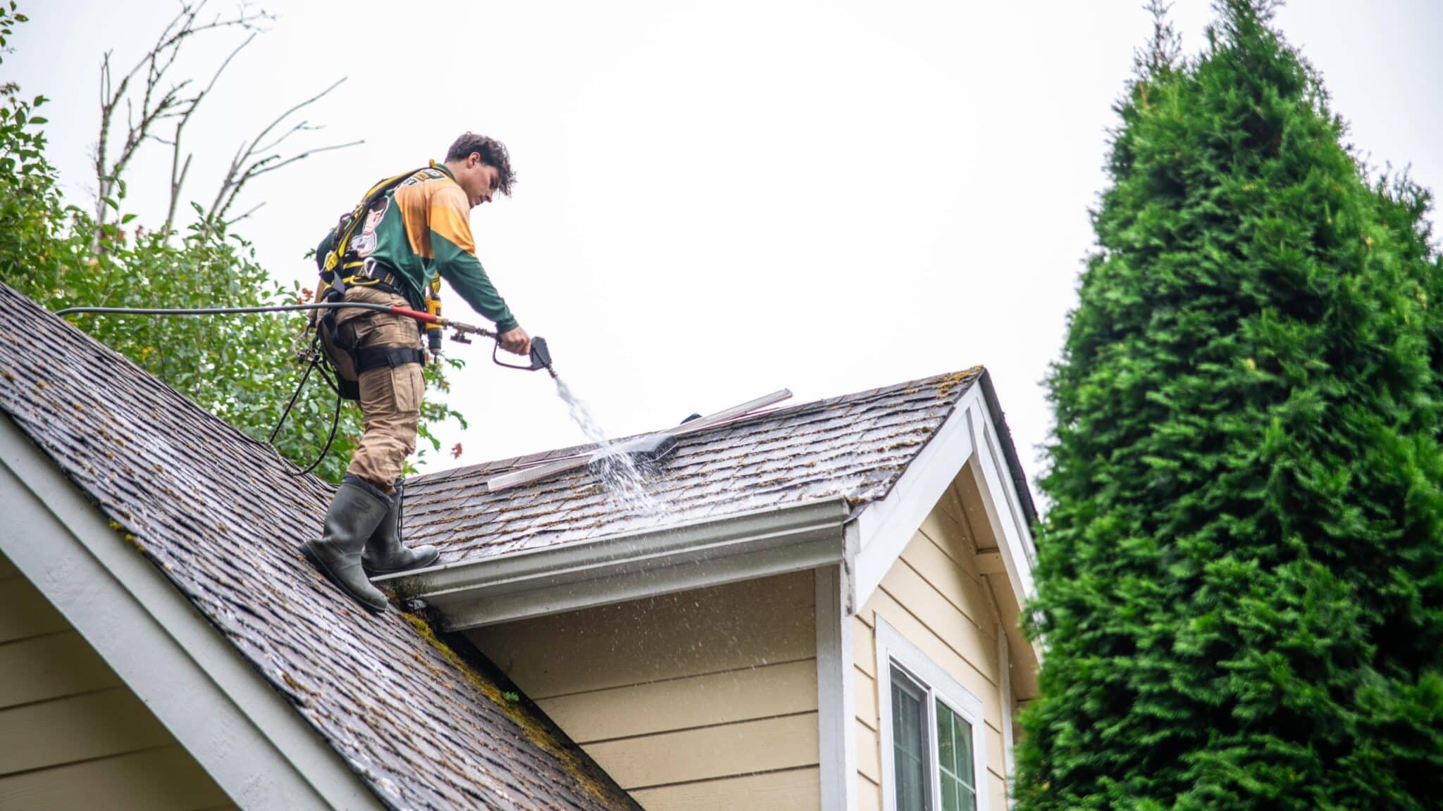Worker on roof removing dirt and debris with a garden hose.