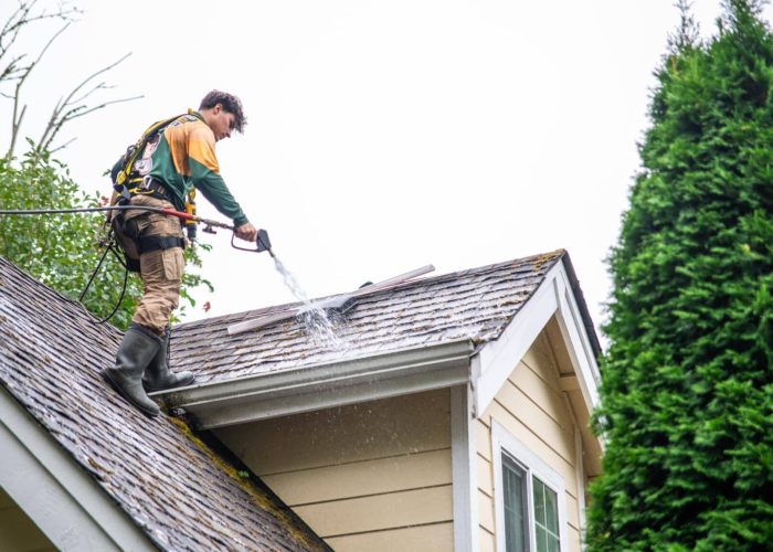 Worker on roof removing dirt and debris with a garden hose.